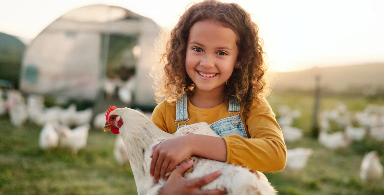 Smiling girl in overalls holding a white chicken on a free-range farm