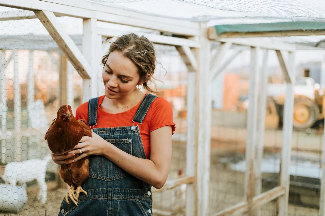 Woman in denim overalls holding a brown hen beside a backyard chicken coop
